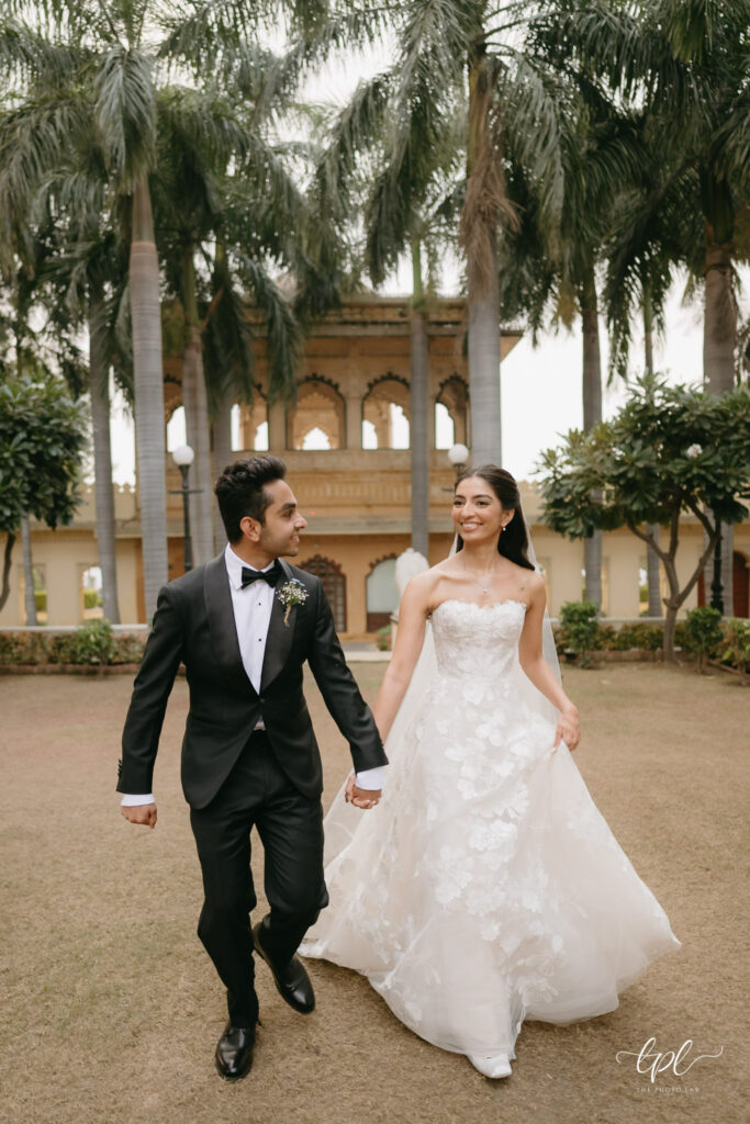 Elegant bride and groom walk hand-in-hand outdoors.