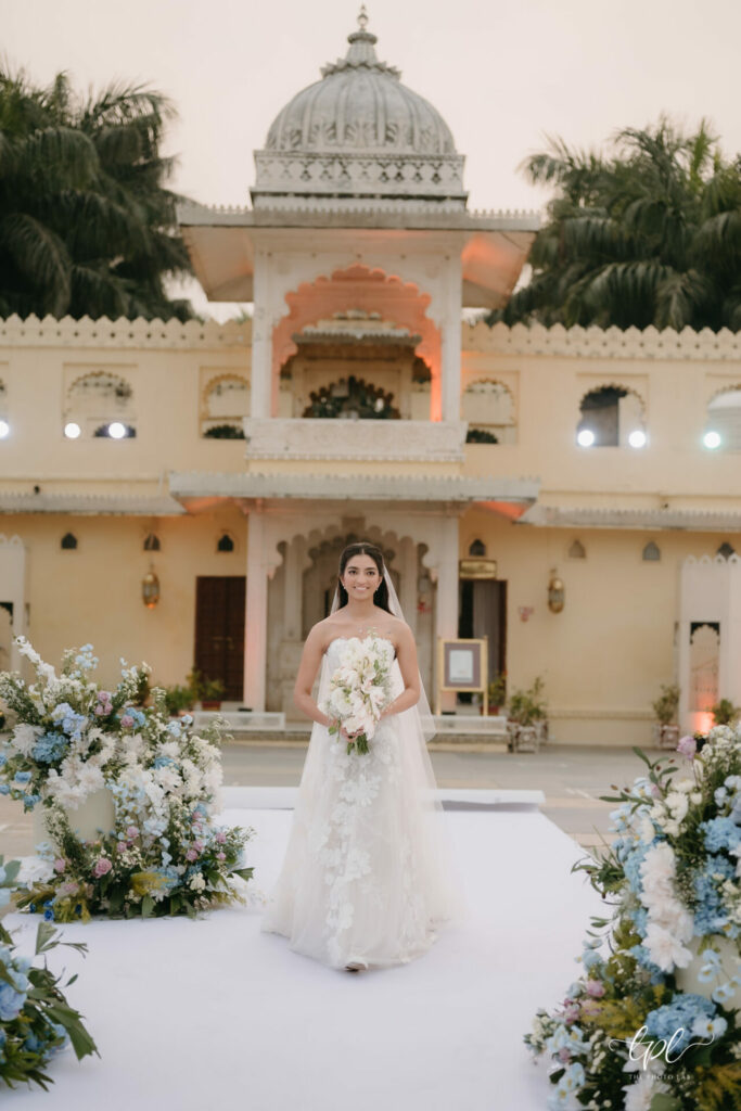 Bride in couture holds white bouquet on floral path at sunset.