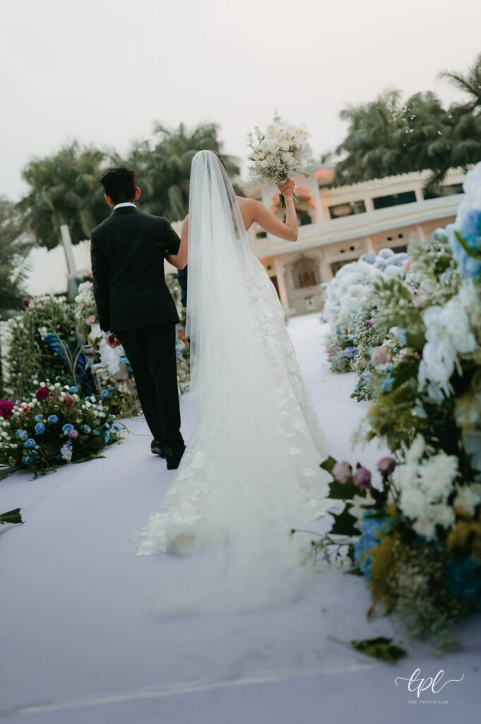 Bride and groom walk outdoor aisle amid pastel flowers.