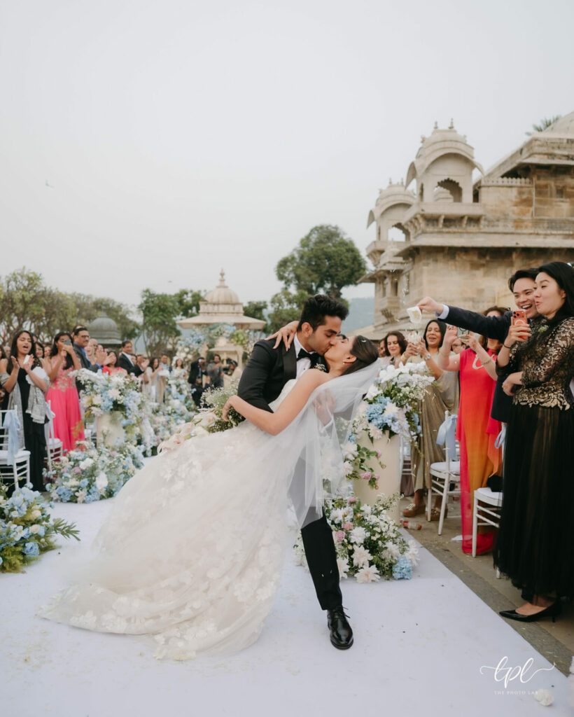 Bride and groom kiss as he dips her outdoors, guests cheer.