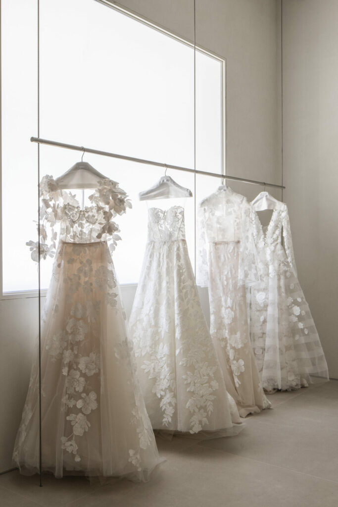 Four white lace wedding dresses hang in soft light.