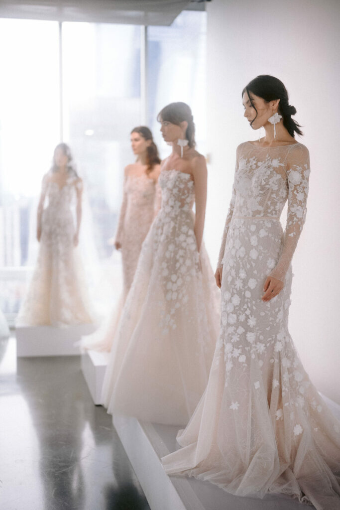 Four women in light lace gowns stand by window.