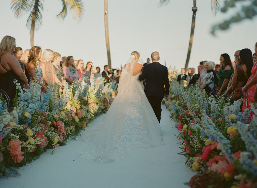 Bride walks flower-lined aisle outdoors amid guests and palms.