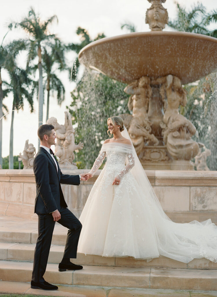 Bride and groom hold hands by ornate fountain.