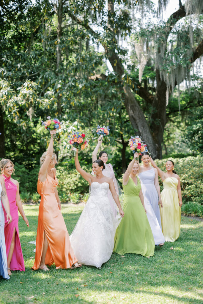 Bride and bridesmaids smiling, bouquets raised on lawn.