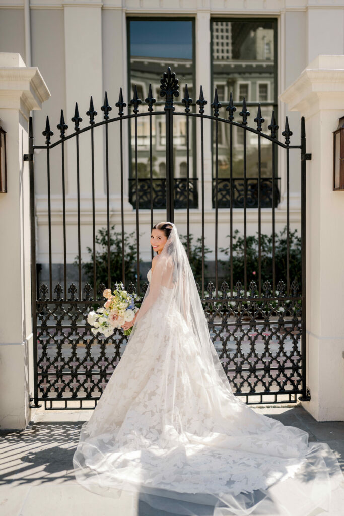 Bride in couture before ornate gates, bouquet in hand.