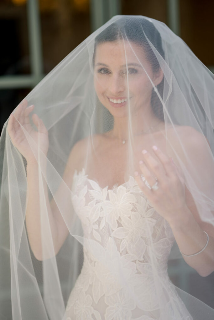 Bride Katya Ruiz smiles in lace gown, holding veil.