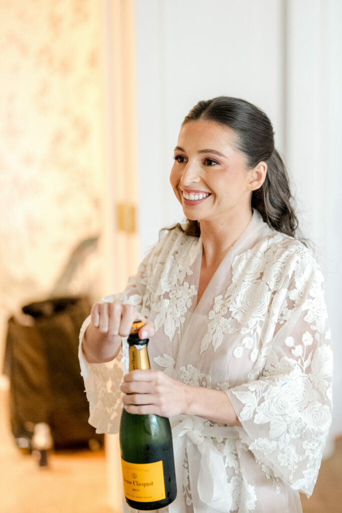 Woman in lace robe opens Veuve Clicquot, beaming.