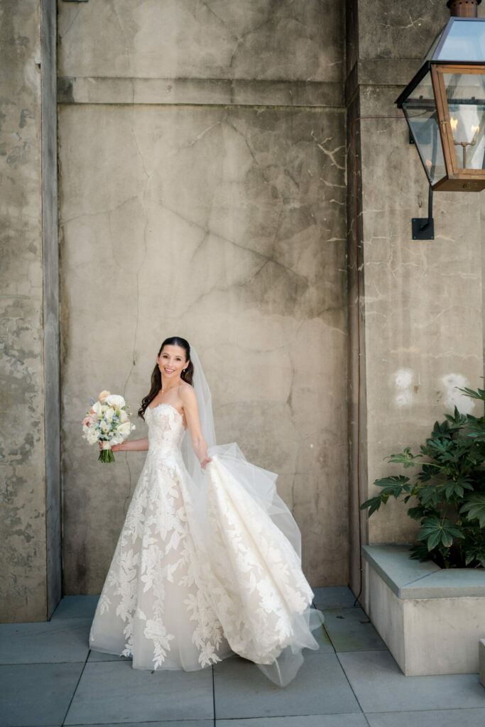 Katya Ruiz smiles in a strapless lace gown, bouquet in hand, beside a stone wall with greenery and a lantern.