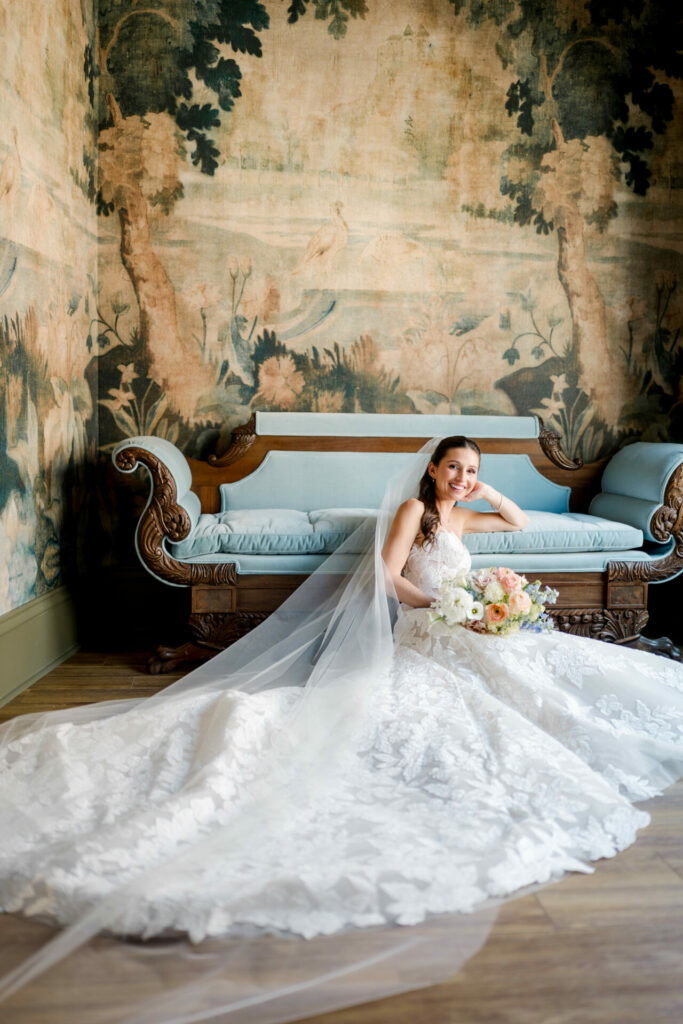 Katya Ruiz sits smiling on vintage blue sofa in lace gown.
