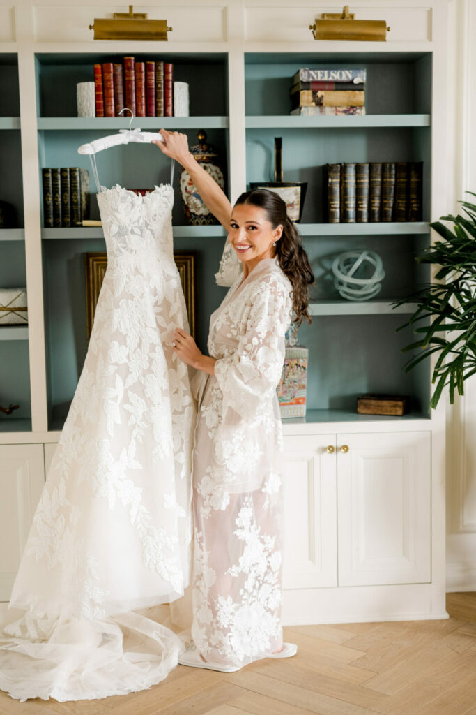 Woman in lace robe holds wedding dress by bookshelf.