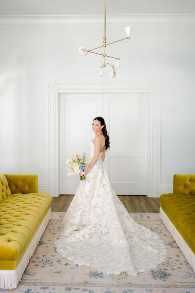 Bride in couture gown holds bouquet between yellow sofas.