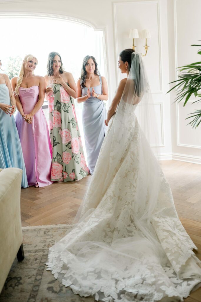 Bride in lace gown admired by pastel-clad bridesmaids.