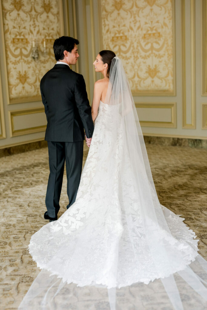 Bride and groom in ornate room, holding hands.