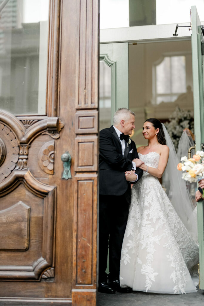 Bride in couture with father, smiling by ornate door.