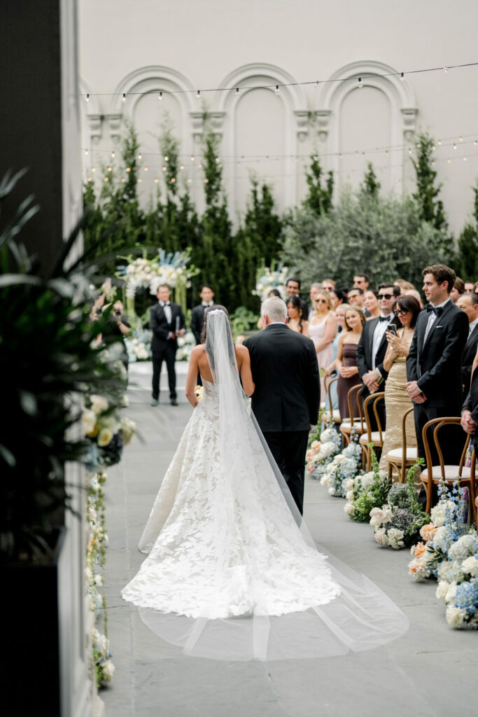 Bride in couture walks outdoor aisle with older man.