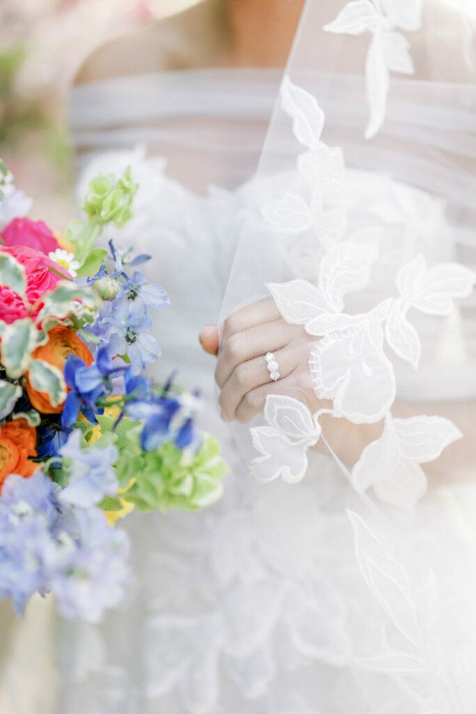 Bride lifts veil, shows ring, holds vibrant bouquet.