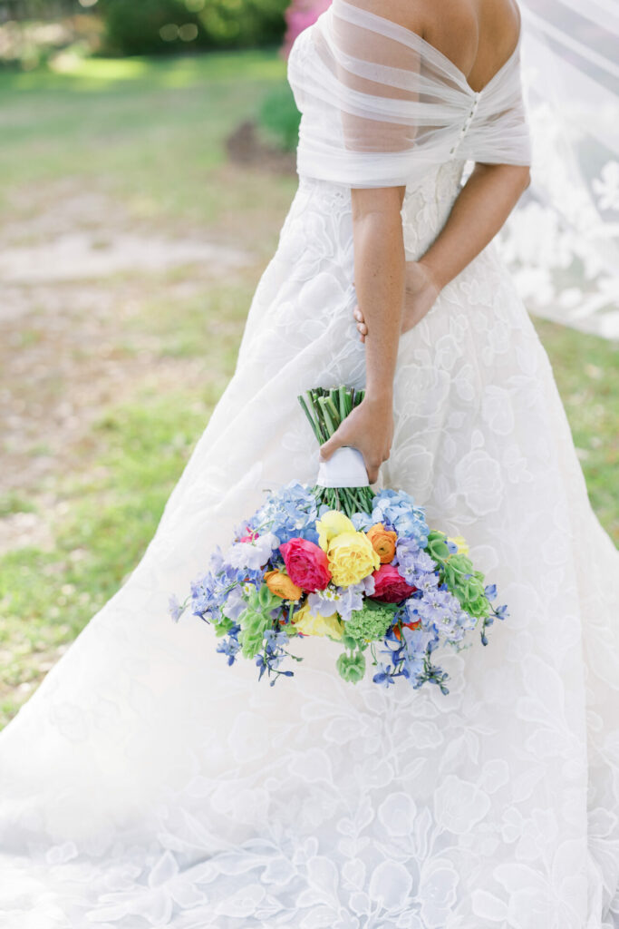 Bride in white gown holding colorful bouquet outdoors.