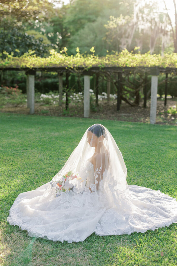 Bride in couture gown sits on sunlit grass.