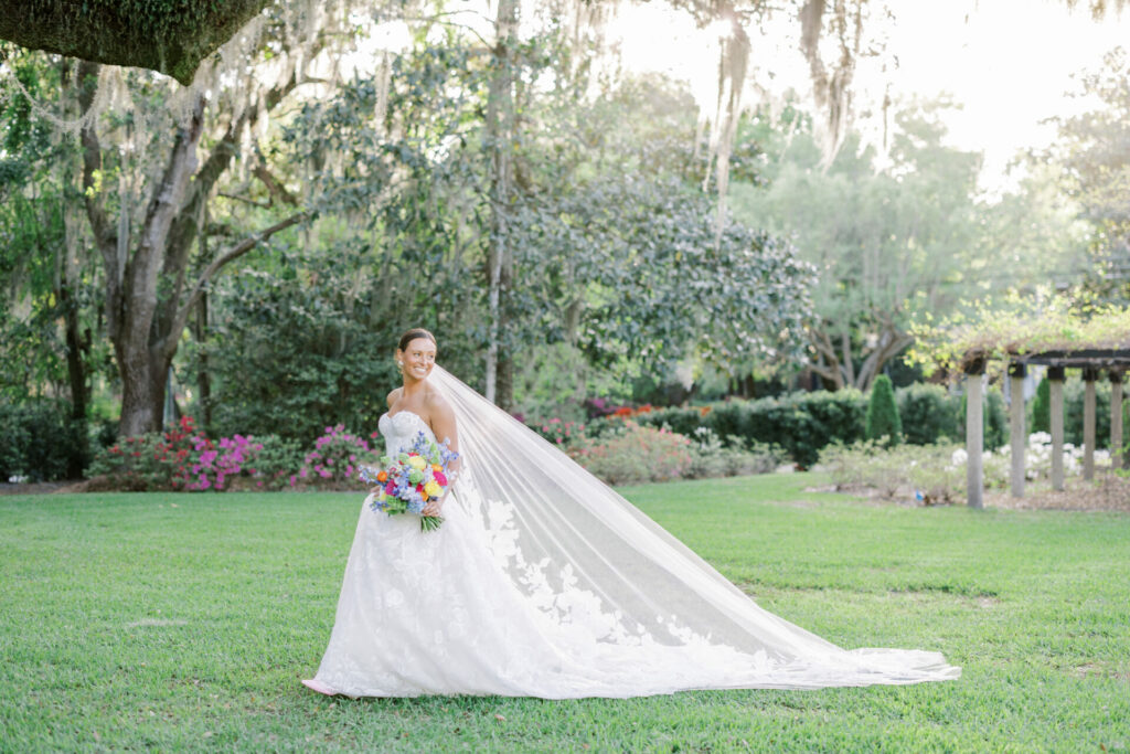 Elegant bride in luxury gown stands in sunlit garden.