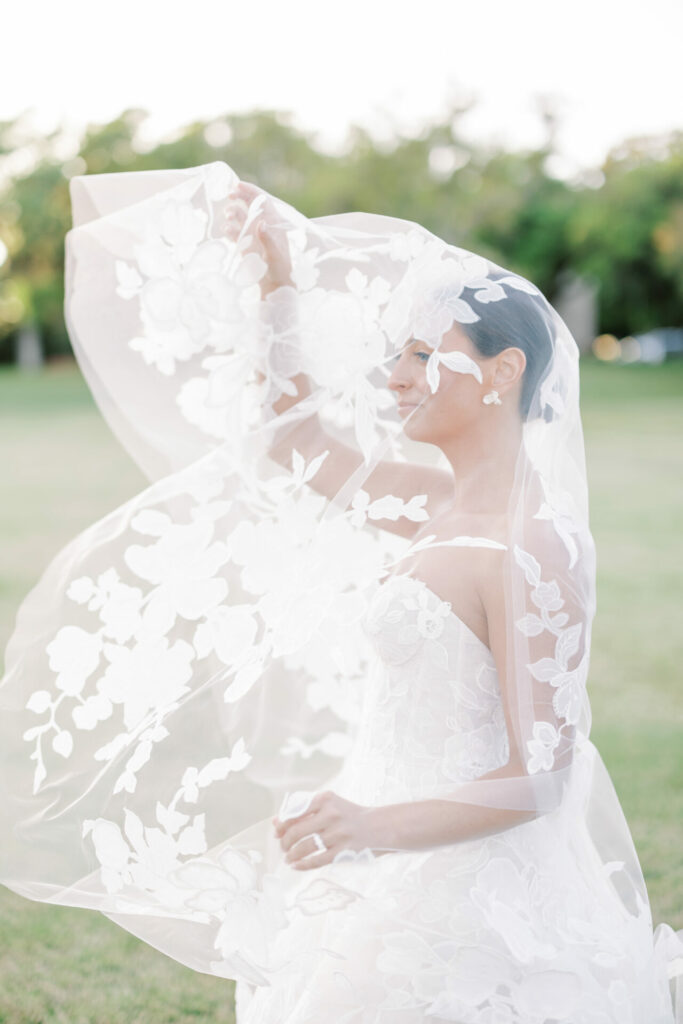 Bride in lace veil smiles on grassy lawn.