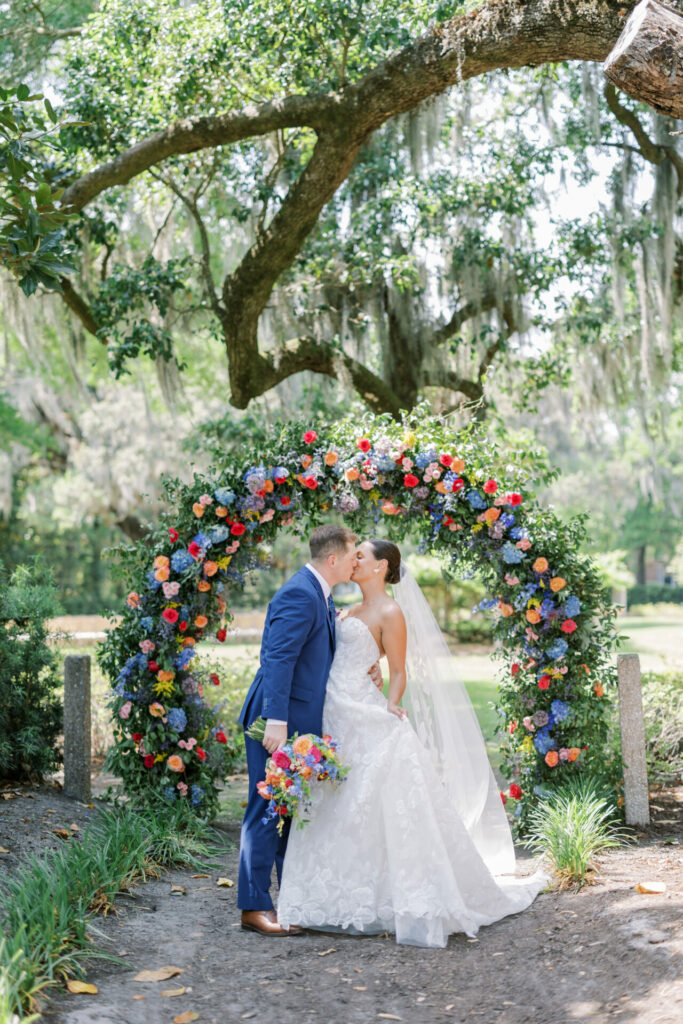 Bride and groom kiss beneath floral arch outdoors.