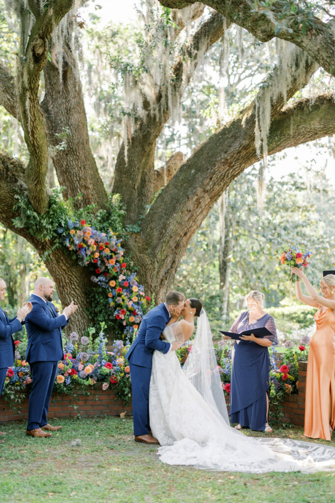 Bride and groom kiss under flower-adorned tree.