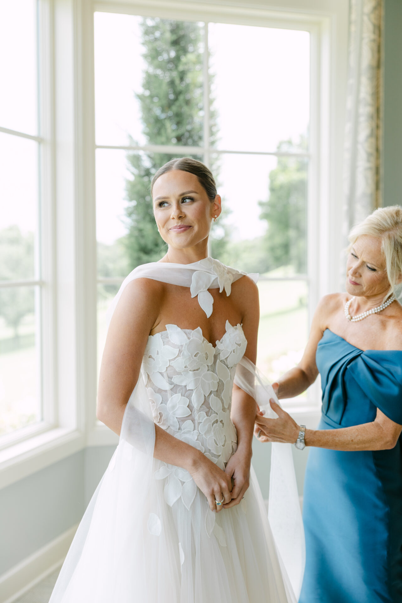 Bride smiles by sunlit window as woman adjusts shawl.