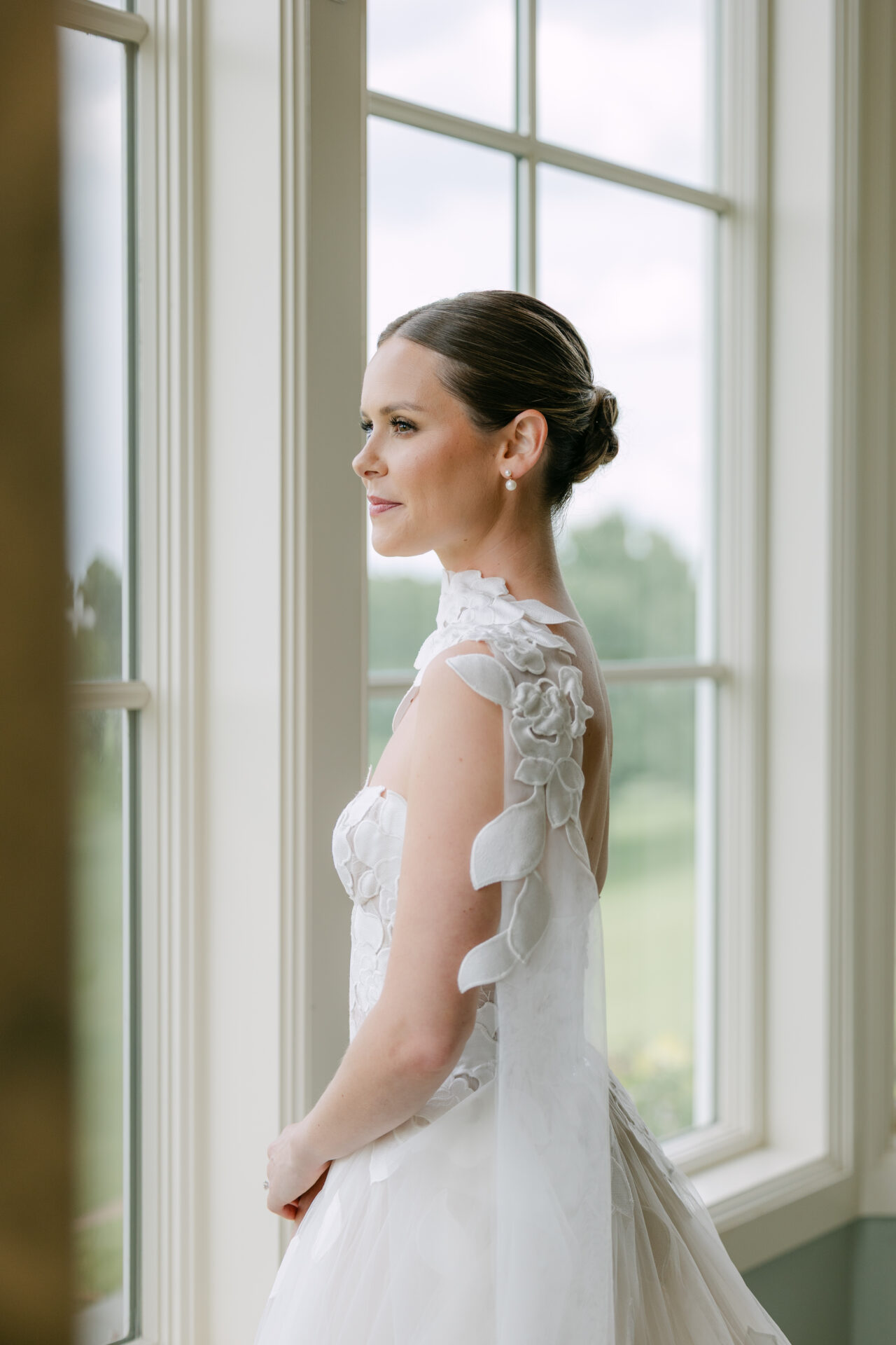 Bride in floral white dress, gazing out window.