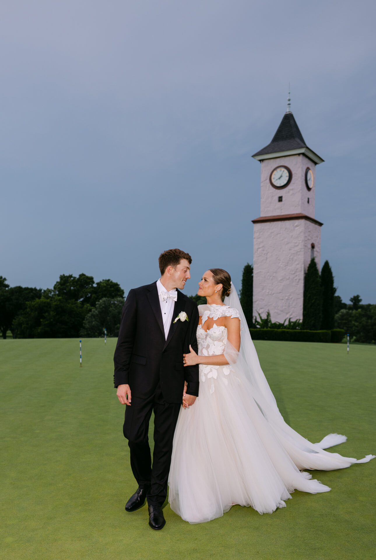 Bride and groom smiling on lawn, clock tower behind.