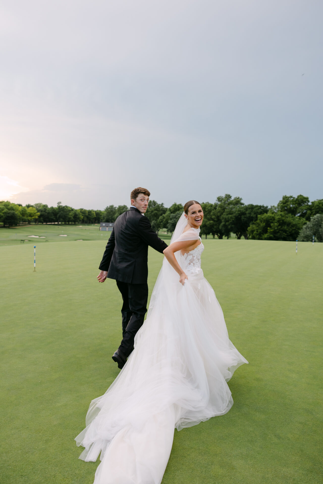 Smiling bride and groom walk hand-in-hand on lawn.