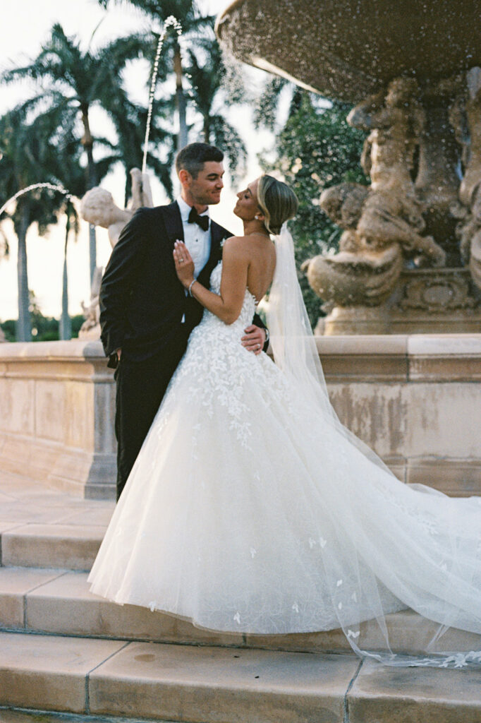 Bride and groom embrace by fountain, palm trees behind.