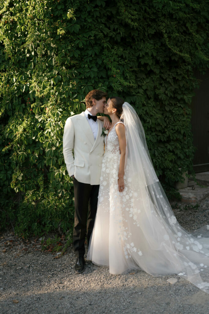 Bride and groom kiss by lush green wall.