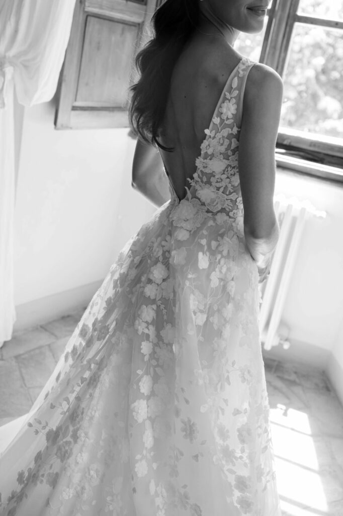 Bride in floral lace gown by sunlit window.