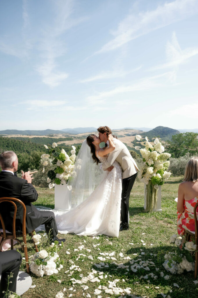 Bride and groom kiss outdoors amid flowers and guests.