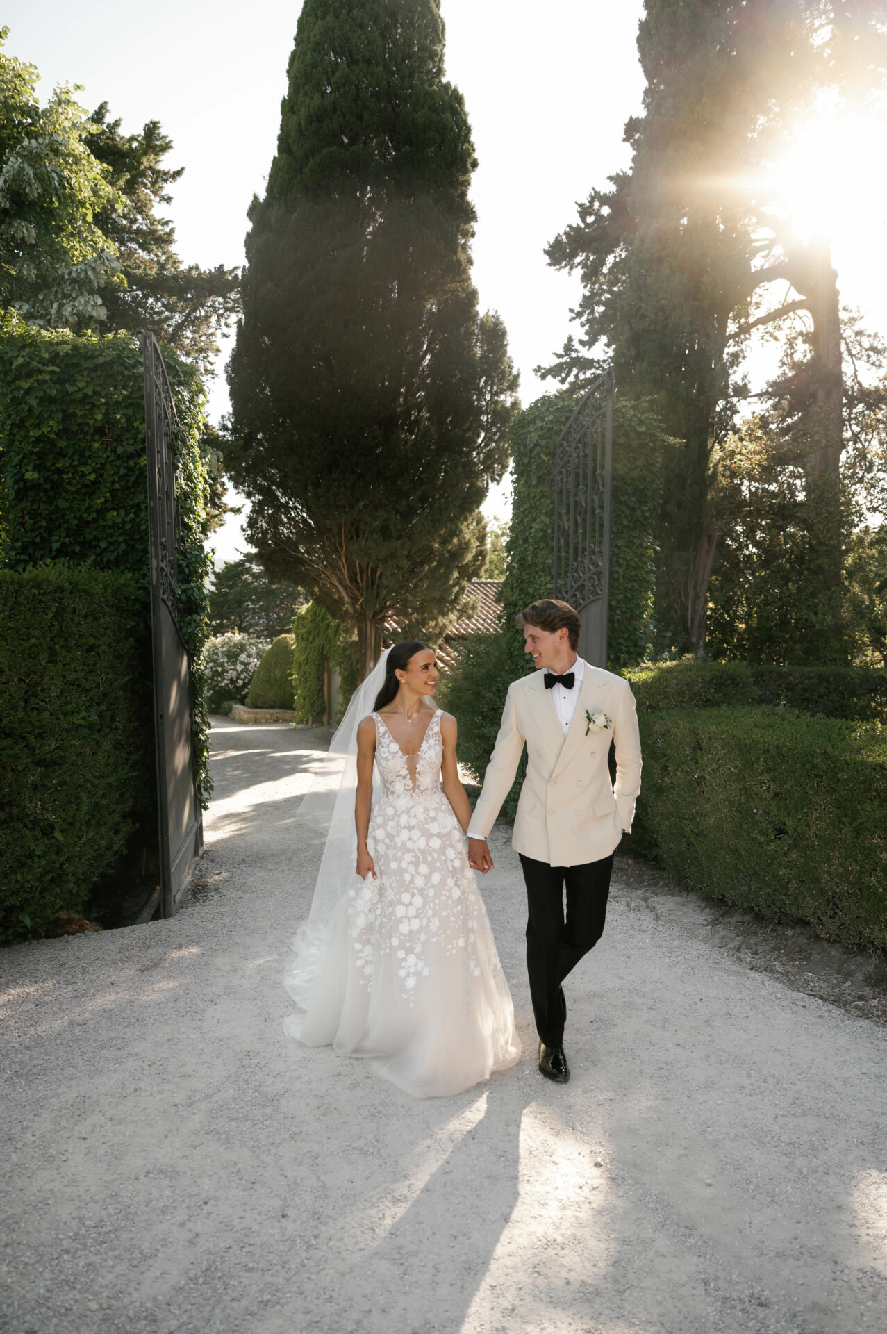 Elegant bride and groom stroll outdoors, sunlit greenery.