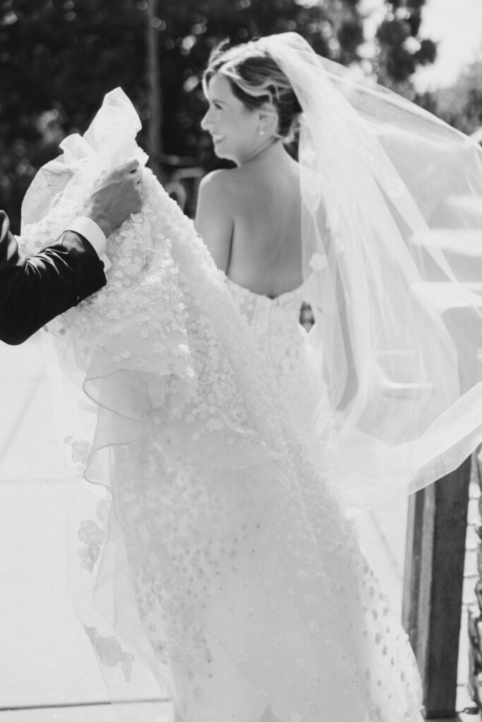 Bride in elegant gown smiles outdoors, veil trailing.