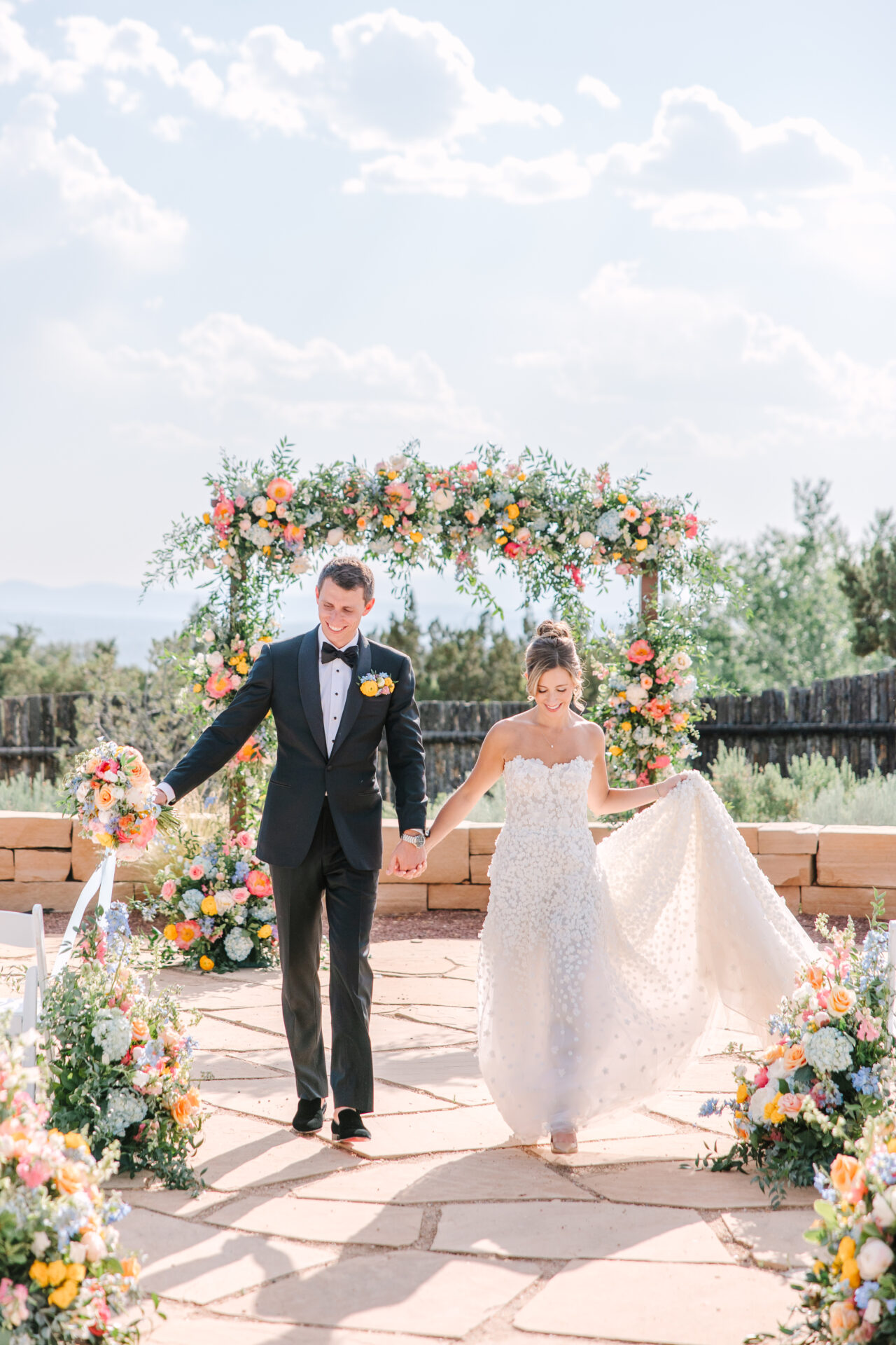 Bride and groom smiling under floral arch outdoors.