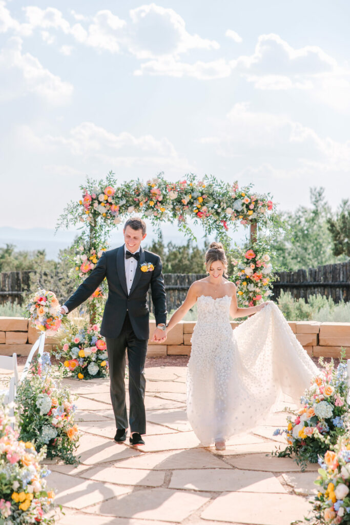 Bride and groom walk under floral outdoor arch.