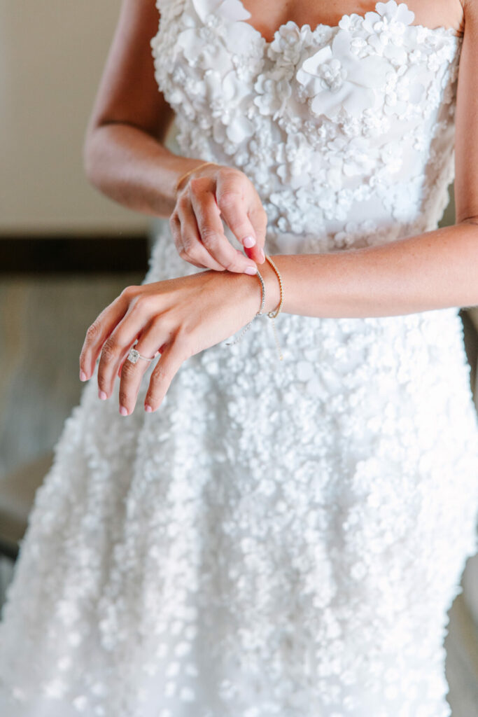 Bride in floral dress fastens bracelet, ring visible.