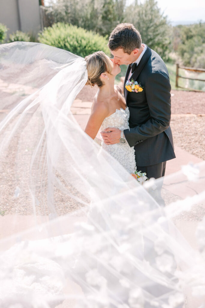 Bride and groom kiss, veil blows in sunlight.