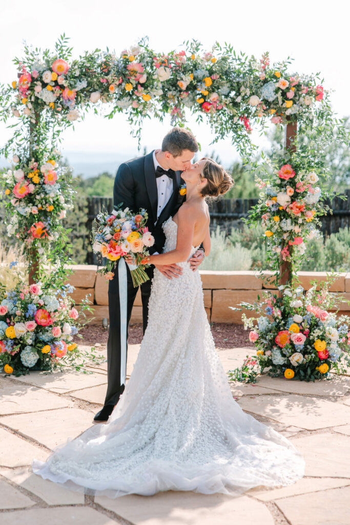 Bride and groom kiss under lush floral arch.