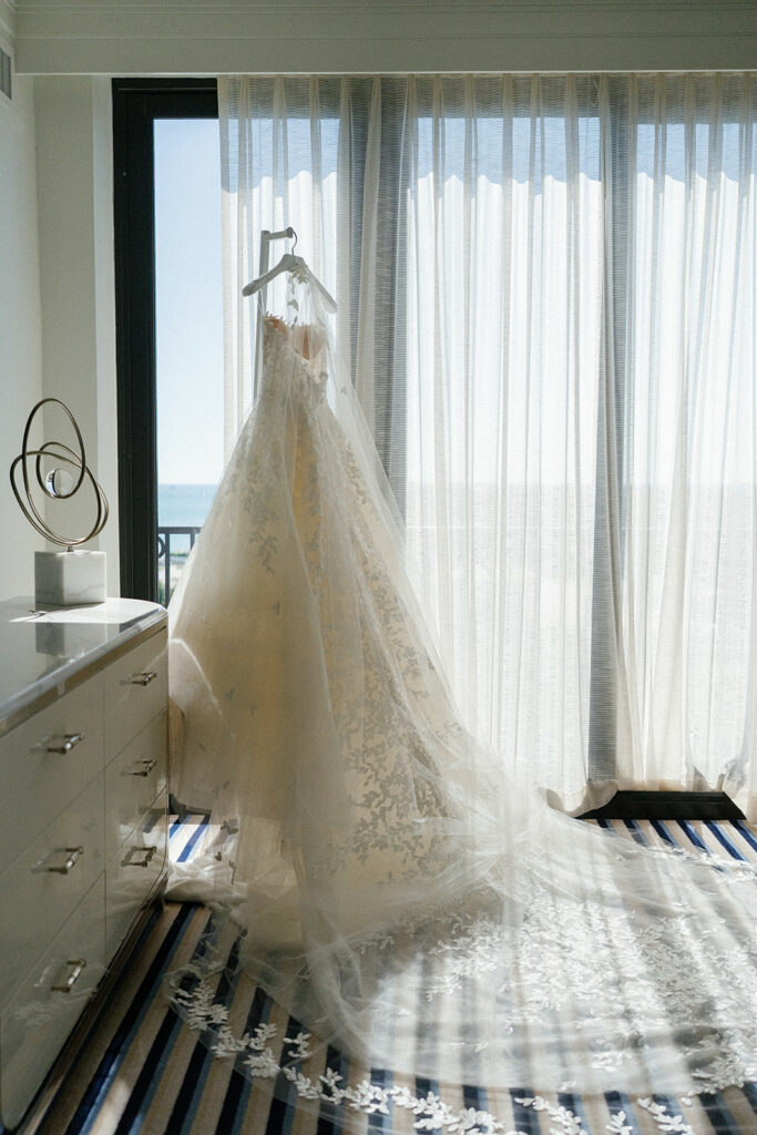 White lace wedding dress hangs by sunny window.
