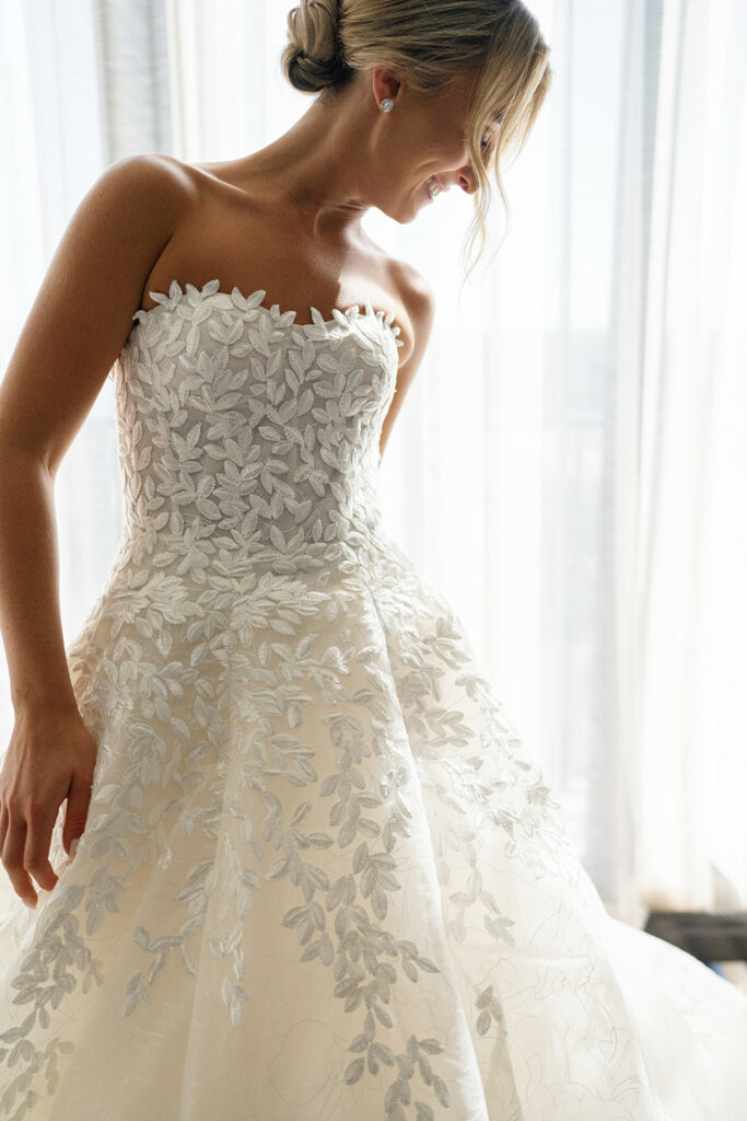 Bride in floral lace dress smiles by window.