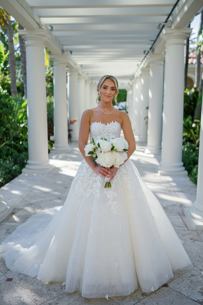 Bride in luxe gown under pergola, holding bouquet.