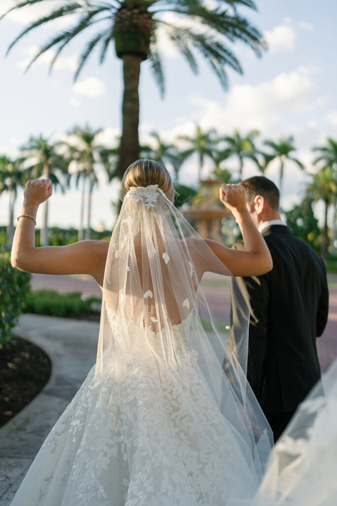 Bride celebrates with groom on sunny palm-lined path.