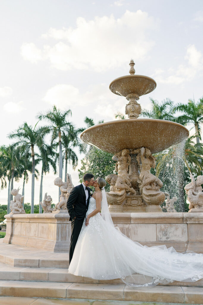 Bride and groom kiss by fountain under palms.
