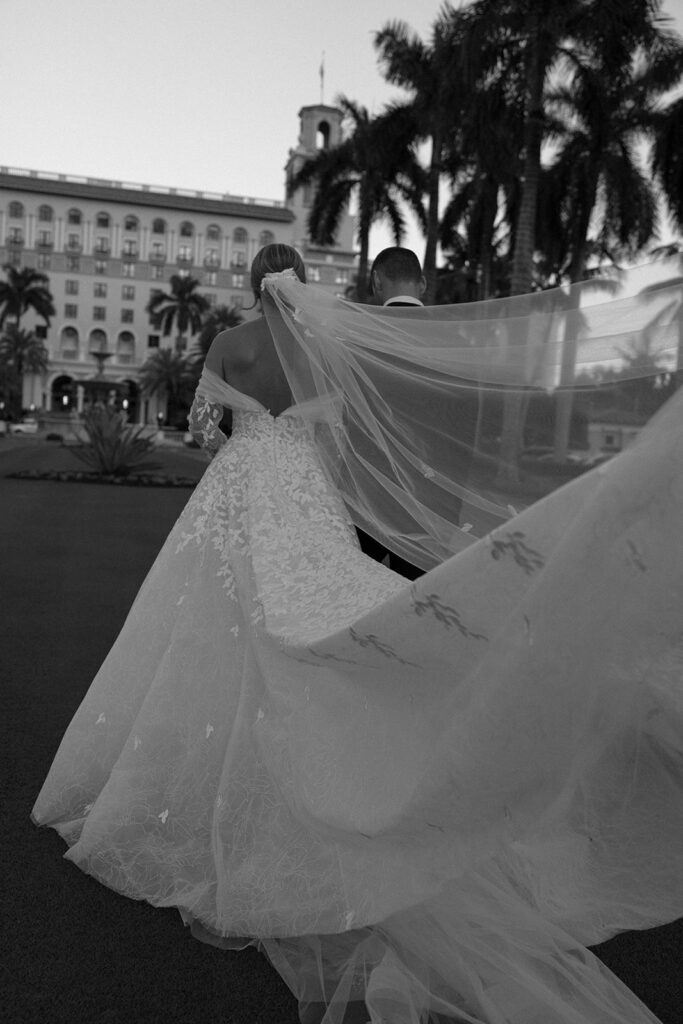 Bride and groom outdoors, flowing veil, elegant backdrop.