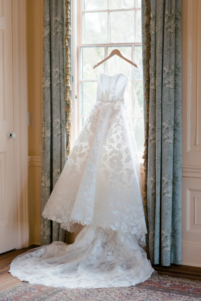 White lace wedding dress hangs in sunlit window.