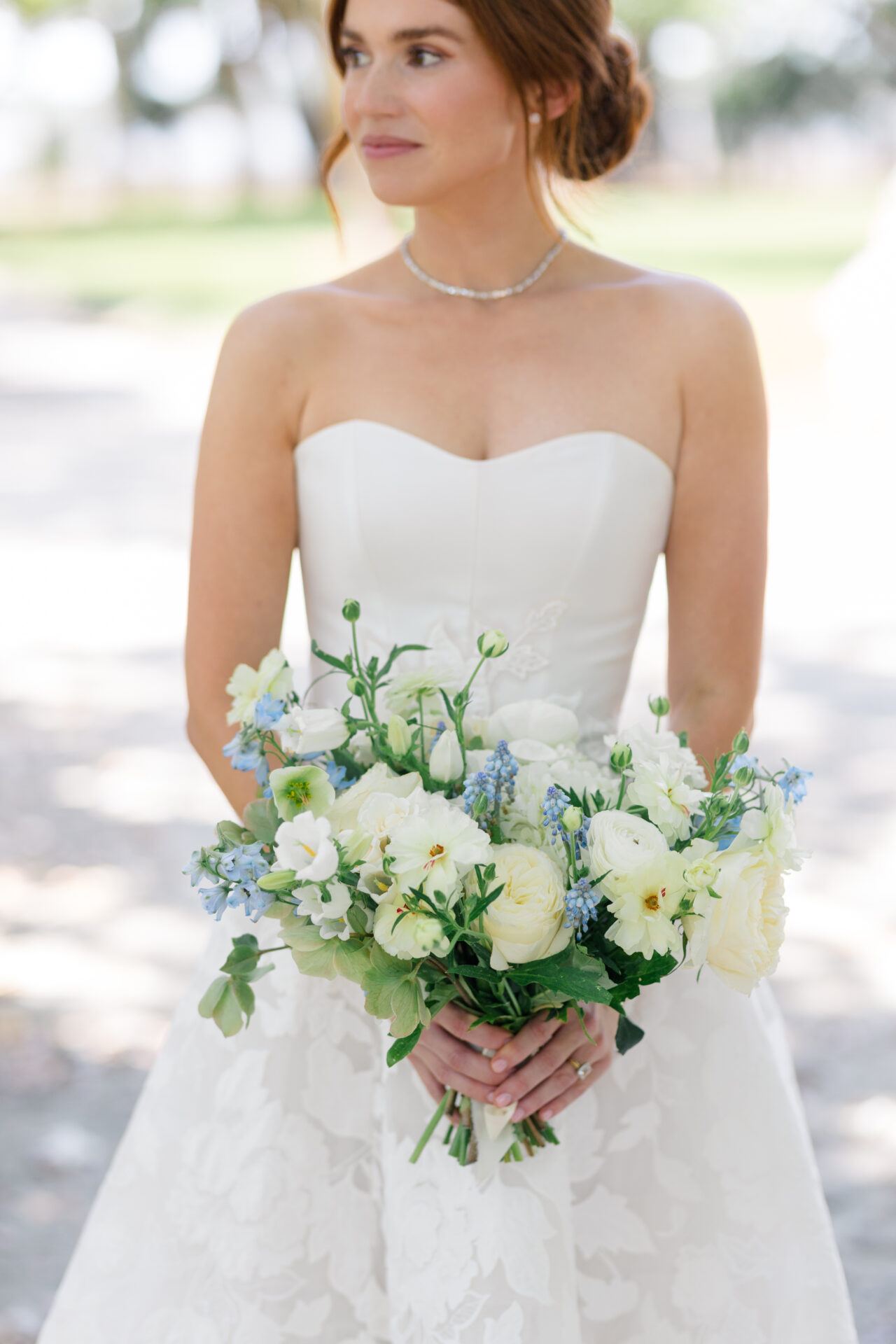 Bride in strapless gown with blue-white bouquet outdoors.